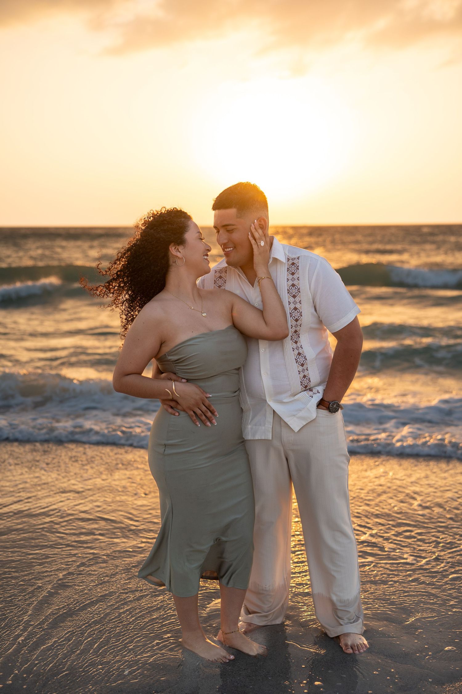 Couple embracing romantically on a beach at golden sunset, waves crashing behind them, wearing elegant casual attire.