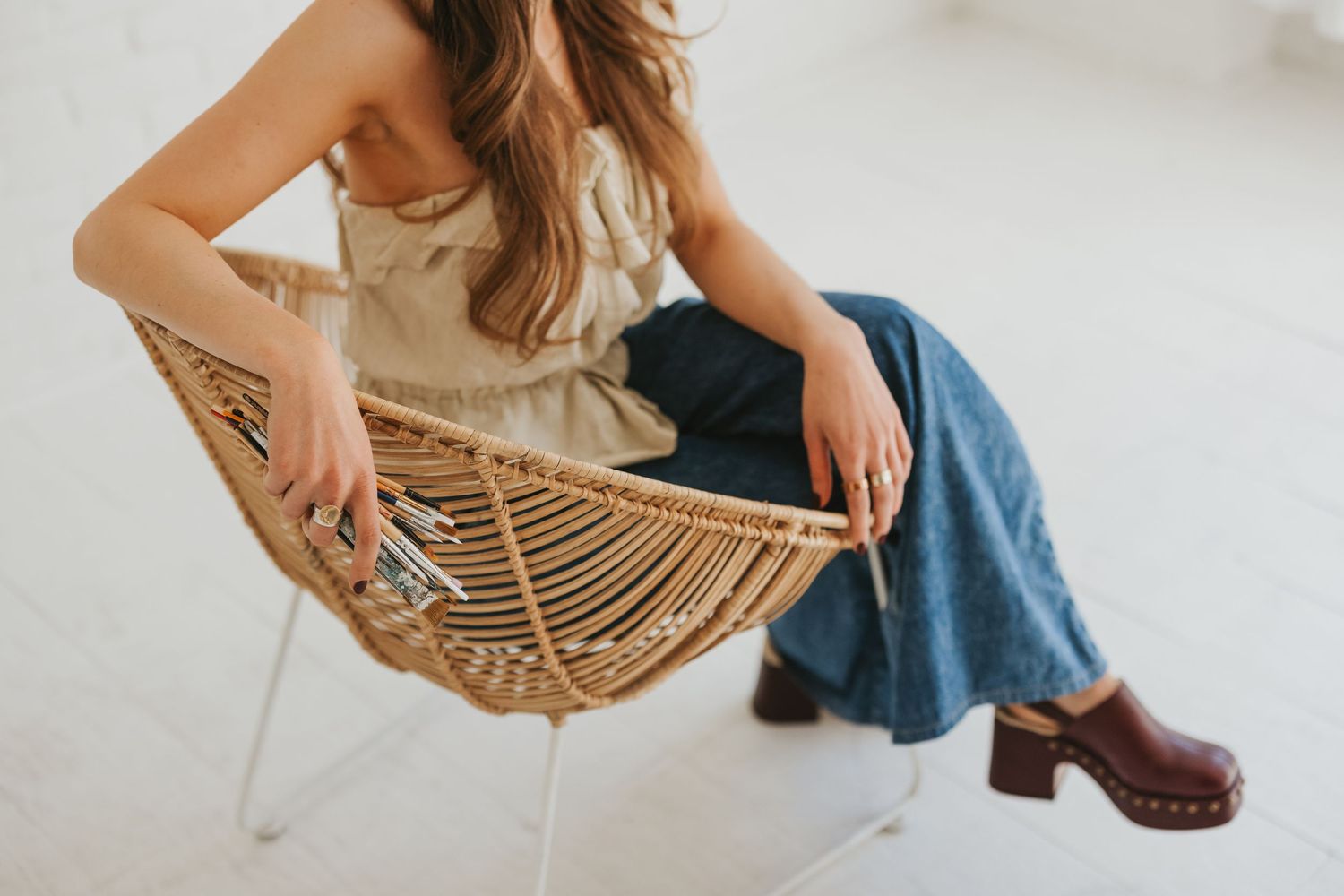 Woman in linen top, blue jeans, and brown clogs sitting in a wicker chair against a white background.