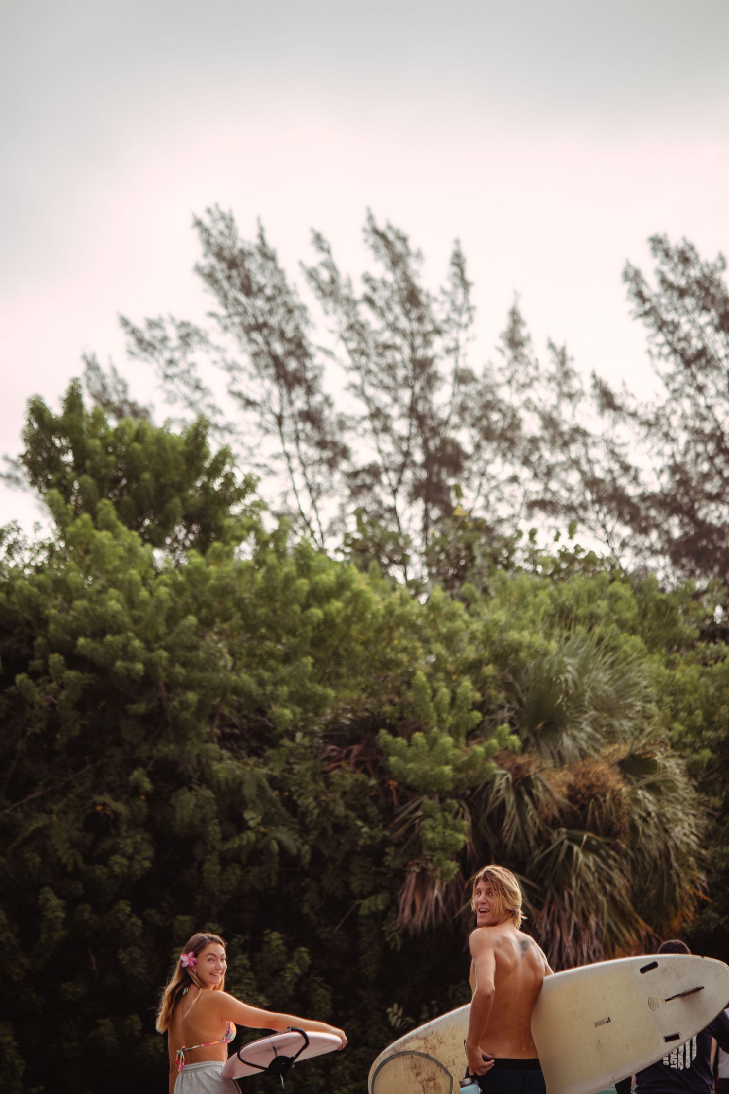 Two surfers carrying surfboards walk near lush green trees on a warm beach day.
