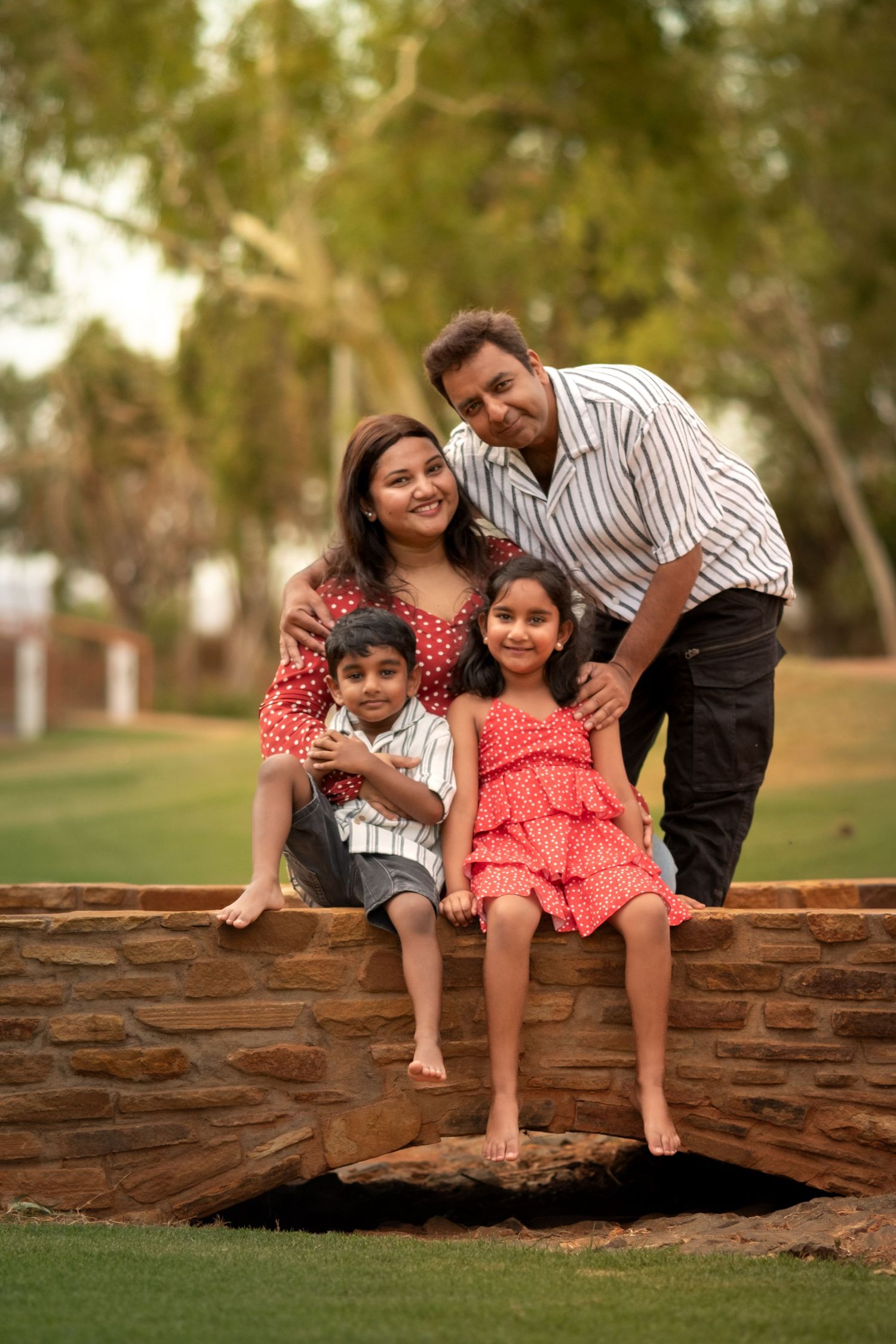 Happy Indian family of four posing outdoors on stone bridge, parents with young son and daughter in red outfits.