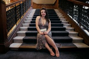 Woman in elegant sequin gown seated on grand staircase with dark wood railings at formal venue.