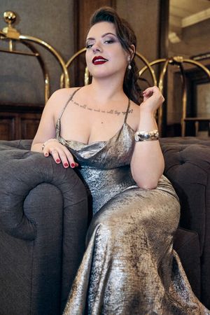 Woman in silver metallic gown seated elegantly in tufted chair with red lips and tattoo.