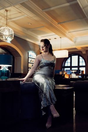 Elegant woman in silver slip dress seated on dark furniture in luxurious hotel lobby.