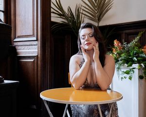 Woman in sequin dress poses at yellow cafe table with tropical flowers and dark wood paneling.