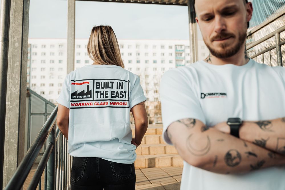 Two models wearing white graphic t-shirts with Built In The East Working Class Heritage print outdoors.
