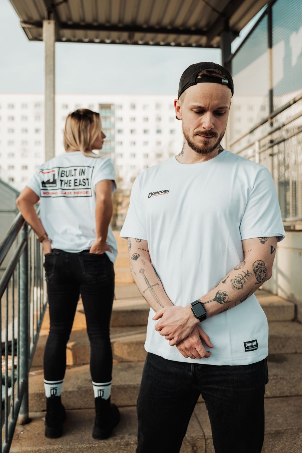 Man with tattoos in white graphic tee and cap poses with woman in matching shirt outdoors.