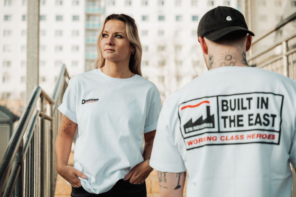Two people wearing white graphic t-shirts, one reading Built In The East Working Class Heroes, standing outdoors.