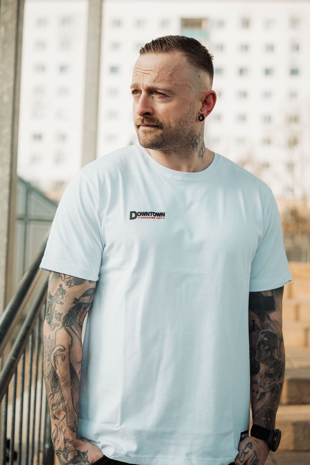 Tattooed man wearing white Downtown branded t-shirt posing outdoors near urban building.