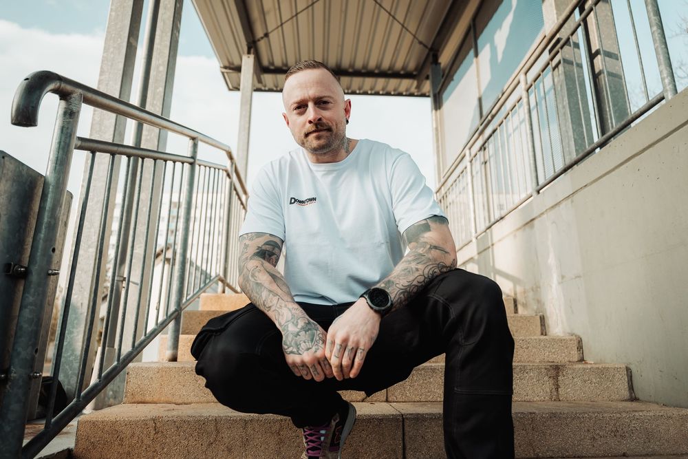 Tattooed bald man in white tee sits on concrete steps outdoors.
