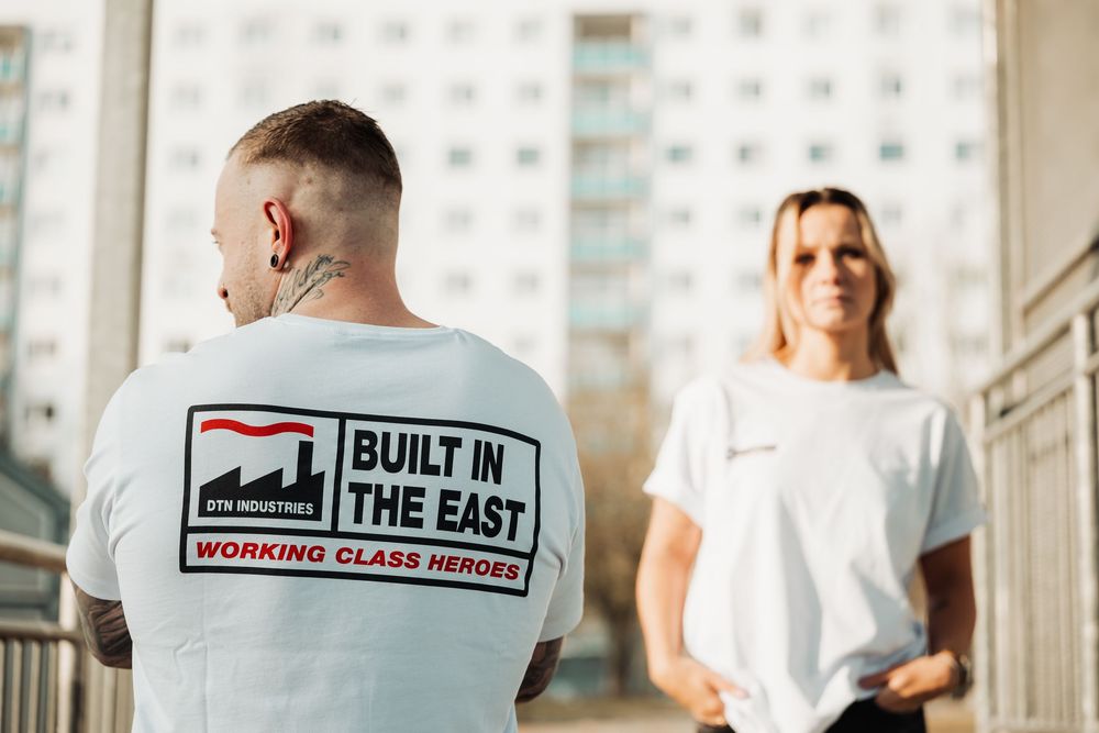 Two people wearing white graphic t-shirts with Built In The East Working Class Heroes print outdoors.