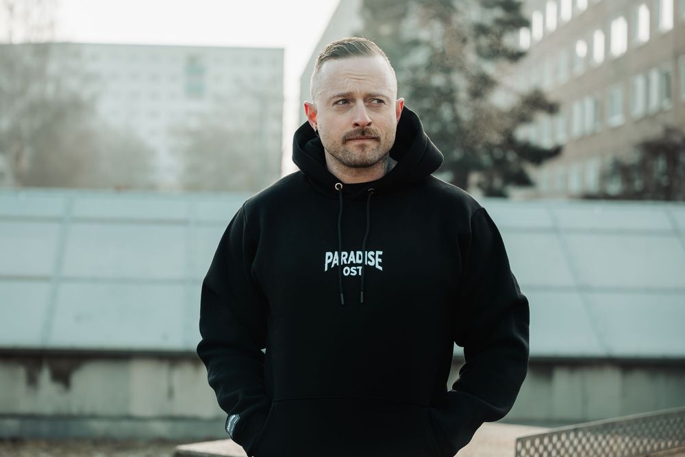 Man wearing black Paradise City hoodie standing outdoors in urban setting.