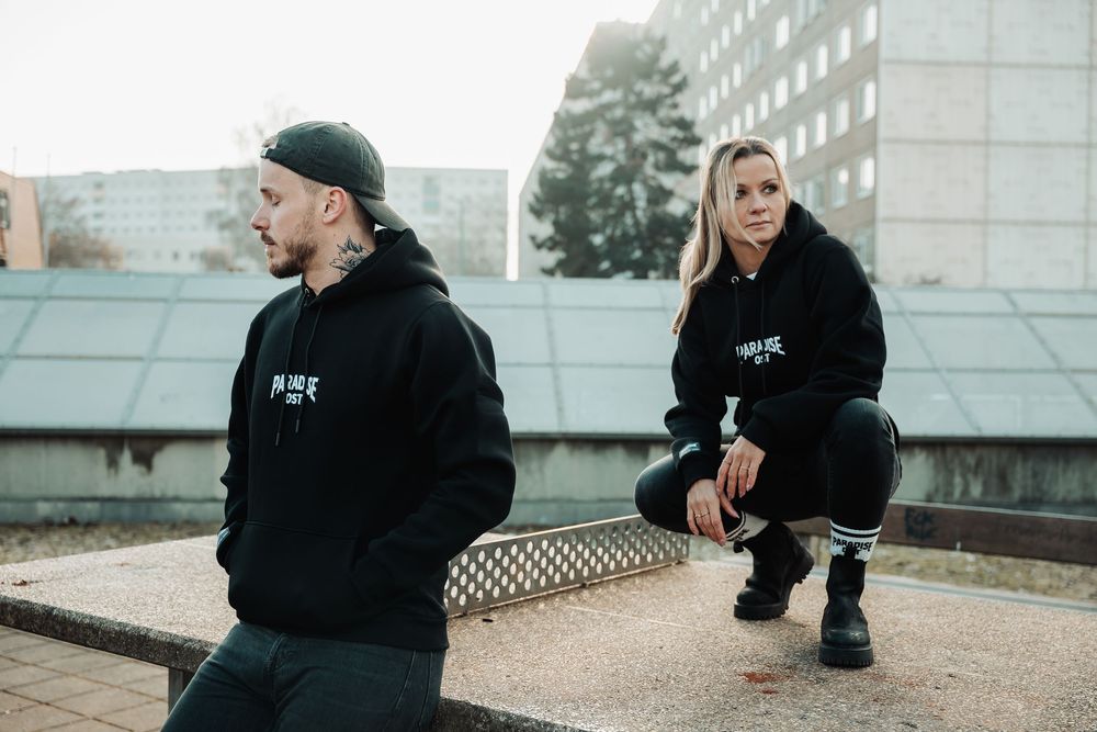 Man and woman in matching black hoodies pose outside urban building.
