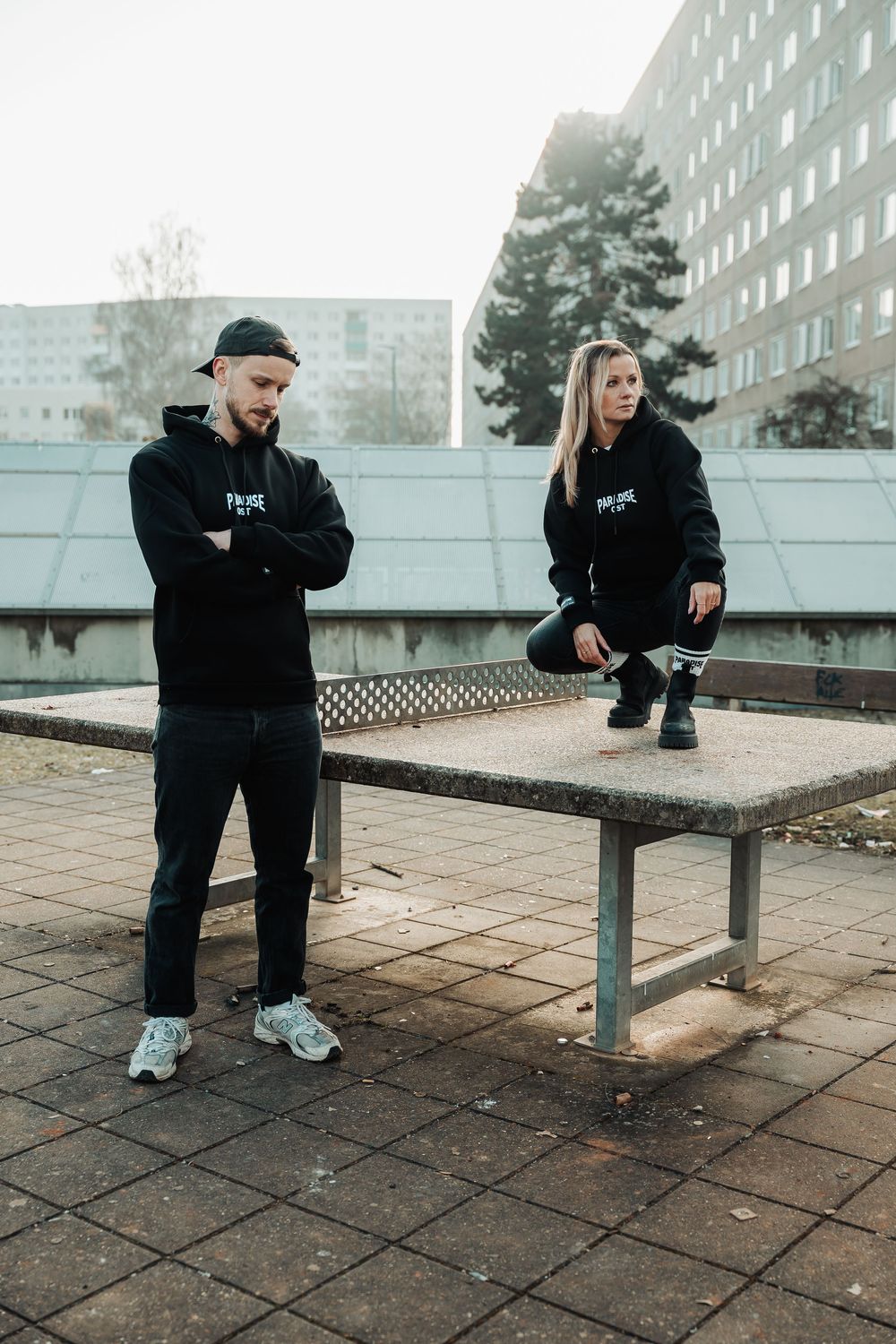 Man and woman wearing matching black Downtown hoodies posing near a concrete table in an urban setting.