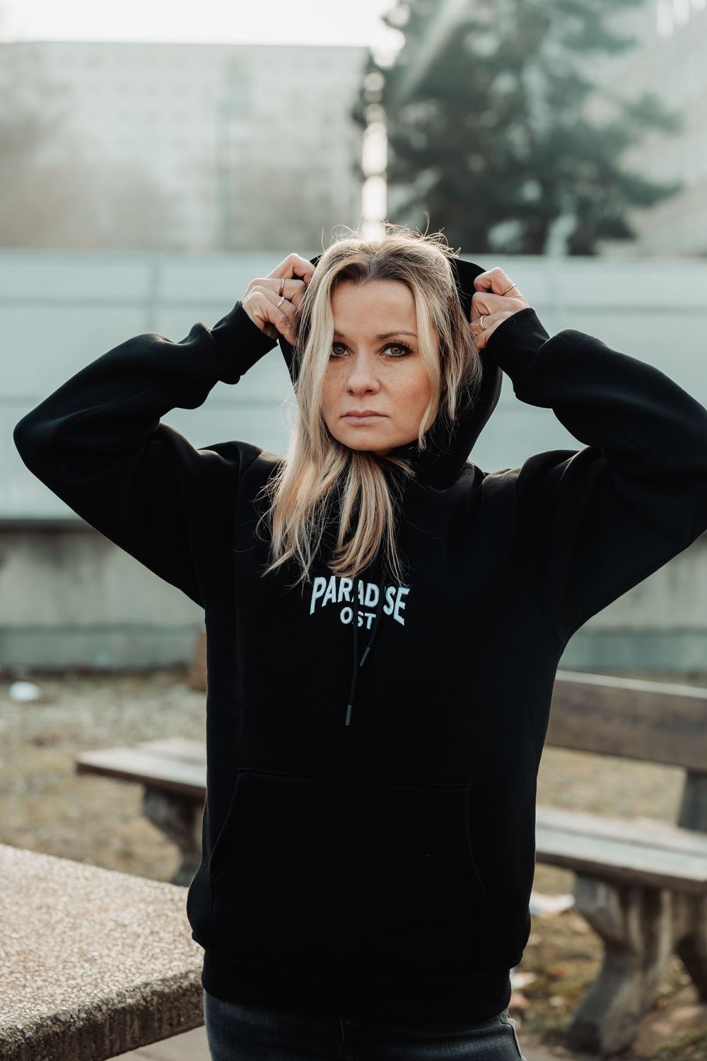 Blonde woman wearing a black Paradise Lost hoodie, pulling up the hood while posing outdoors near benches.
