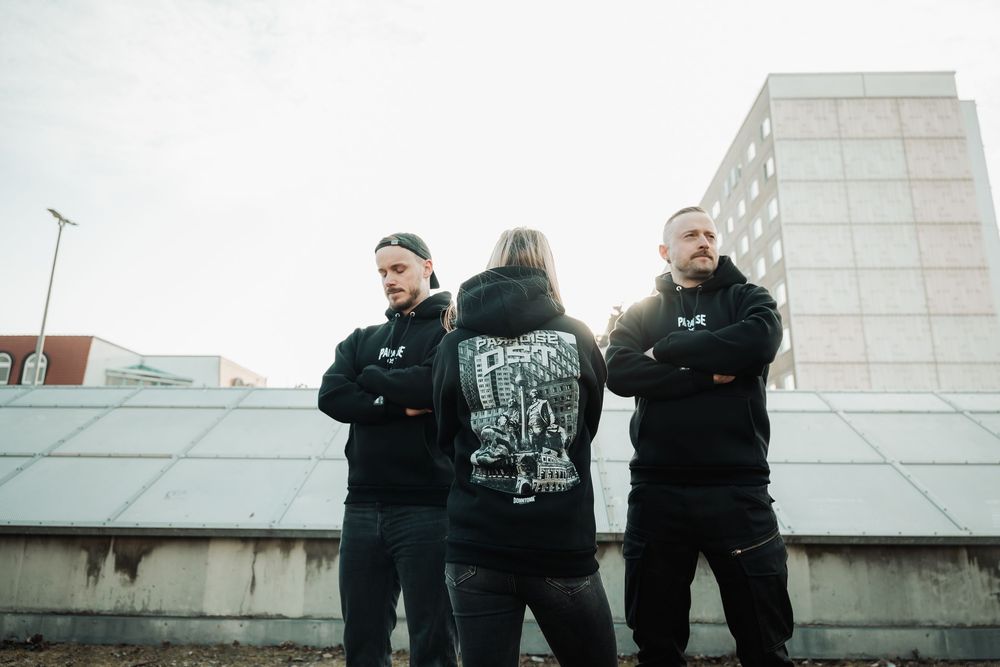 Three people in black hoodies stand on rooftop with urban backdrop.