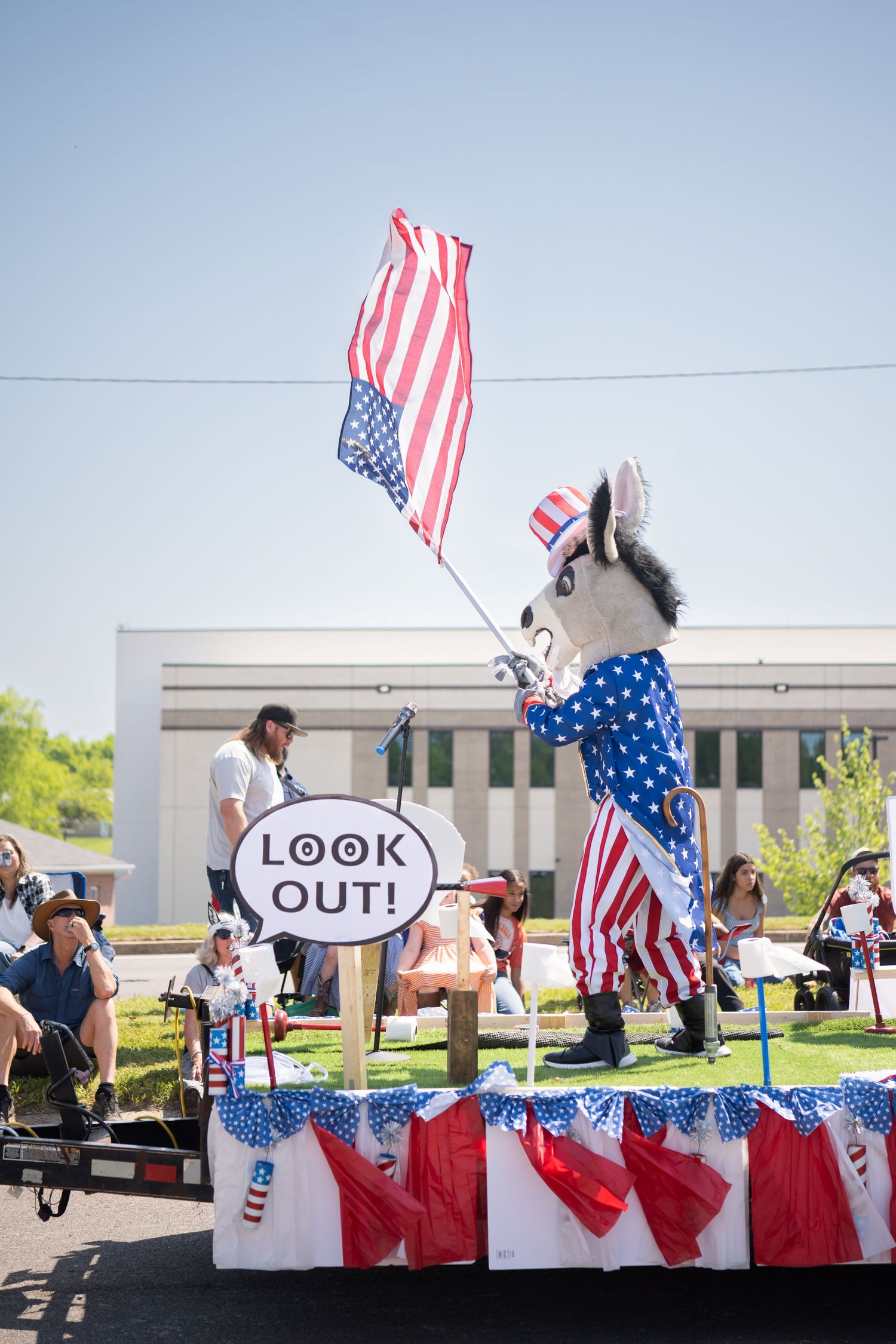 Patriotic mascot in Uncle Sam costume waves American flag on a parade float with a Look Out sign.