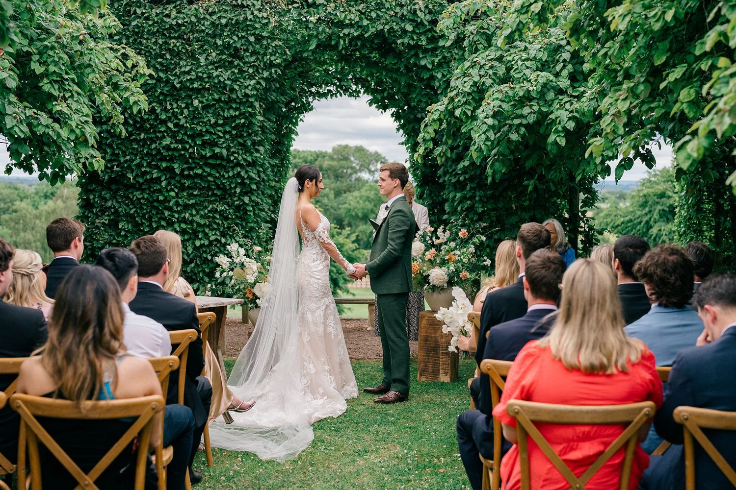 Bride and groom exchange vows under a lush green ivy arch at an outdoor garden wedding ceremony with seated guests.