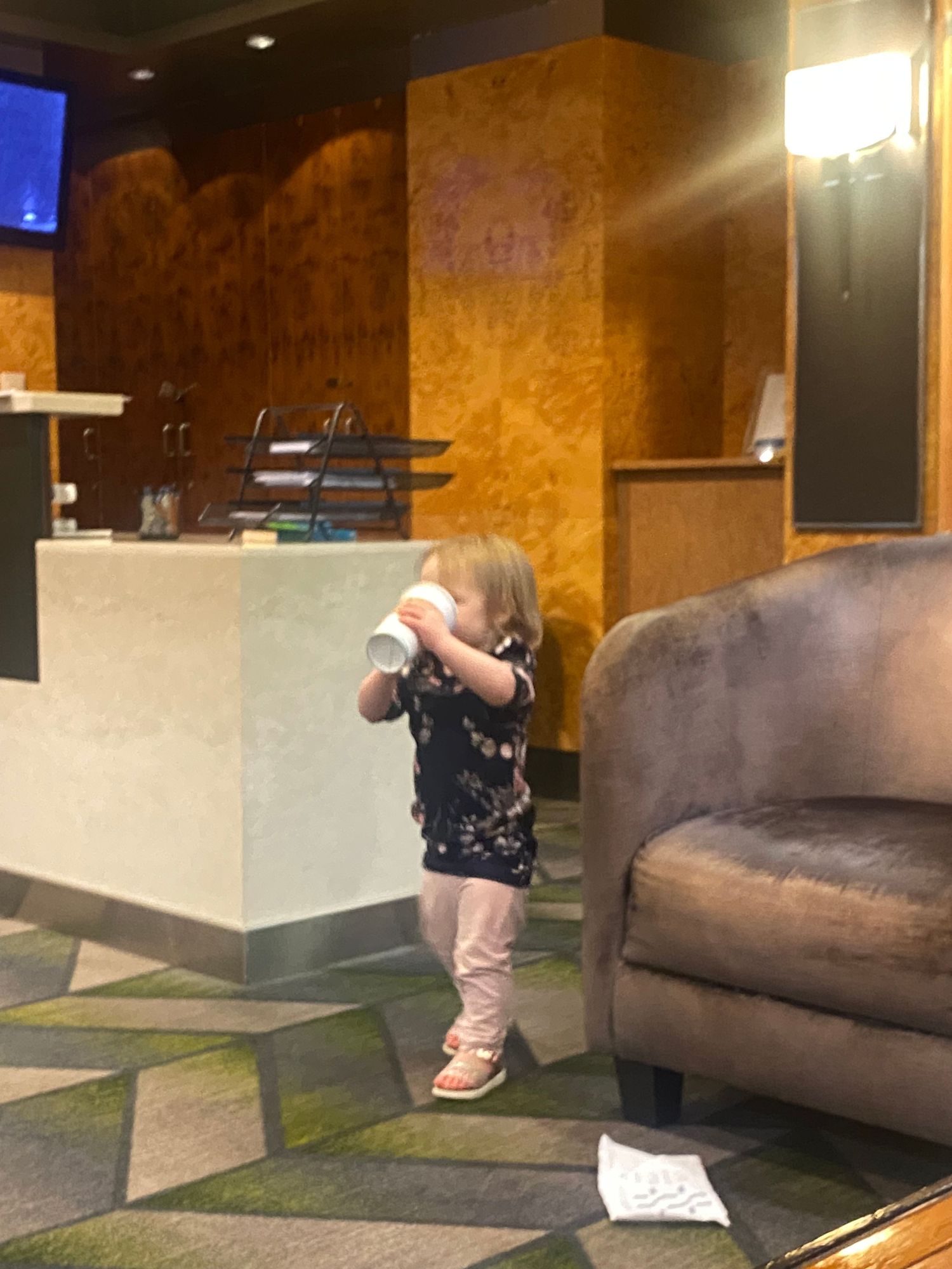 Toddler girl drinking from a cup in a hotel lobby near a reception desk and brown armchair.