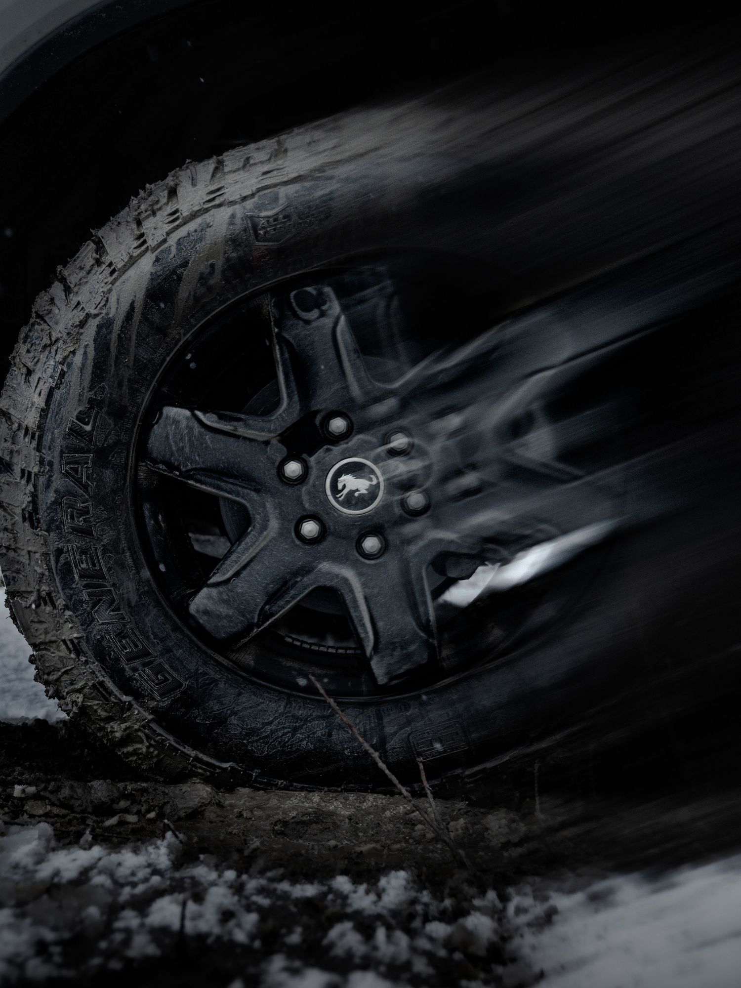 Black alloy wheel with pony emblem spinning on snowy terrain, motion blur effect on dark background.