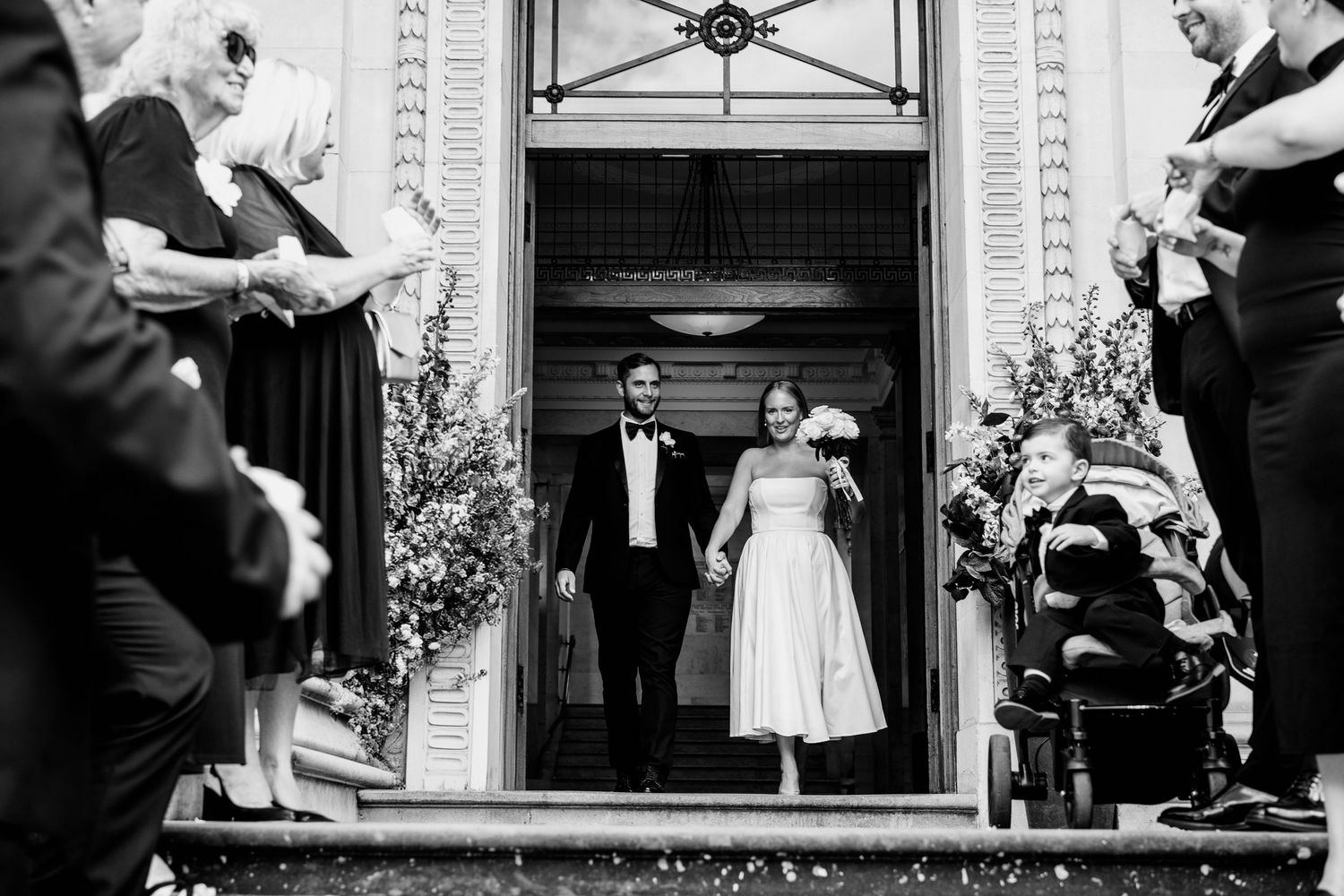 Black and white photo of a bride and groom exiting a grand building entrance, greeted by cheering wedding guests.
