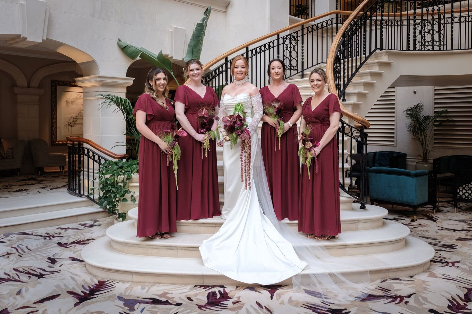 Bride in white gown with bridesmaids in burgundy dresses holding bouquets on elegant staircase landing.