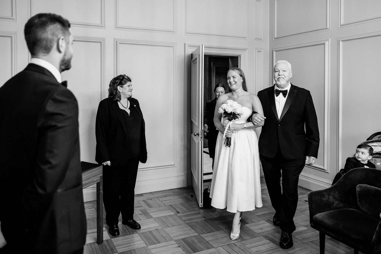 Black and white photo of a bride walking down the aisle with her father at an elegant indoor wedding ceremony.