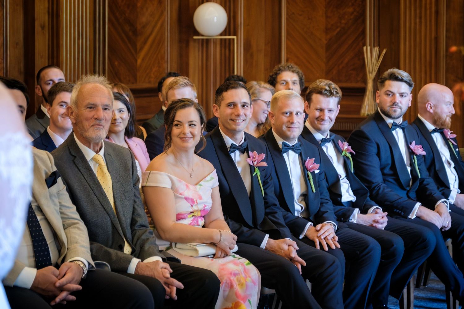 Wedding guests smiling and seated during a ceremony, with groomsmen in navy suits with pink boutonnieres.