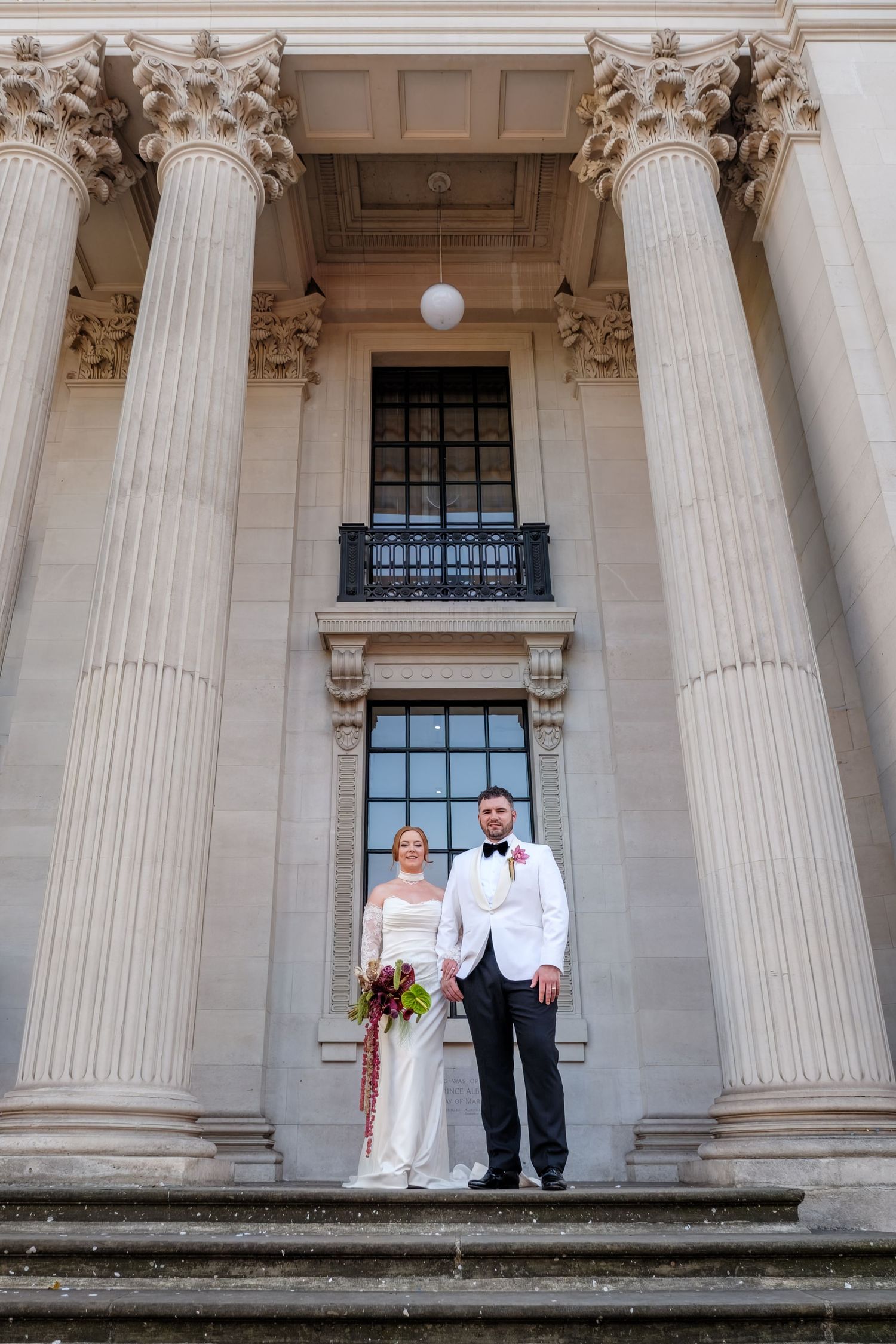 Bride and groom pose on steps of grand neoclassical building with tall Corinthian columns on their wedding day.
