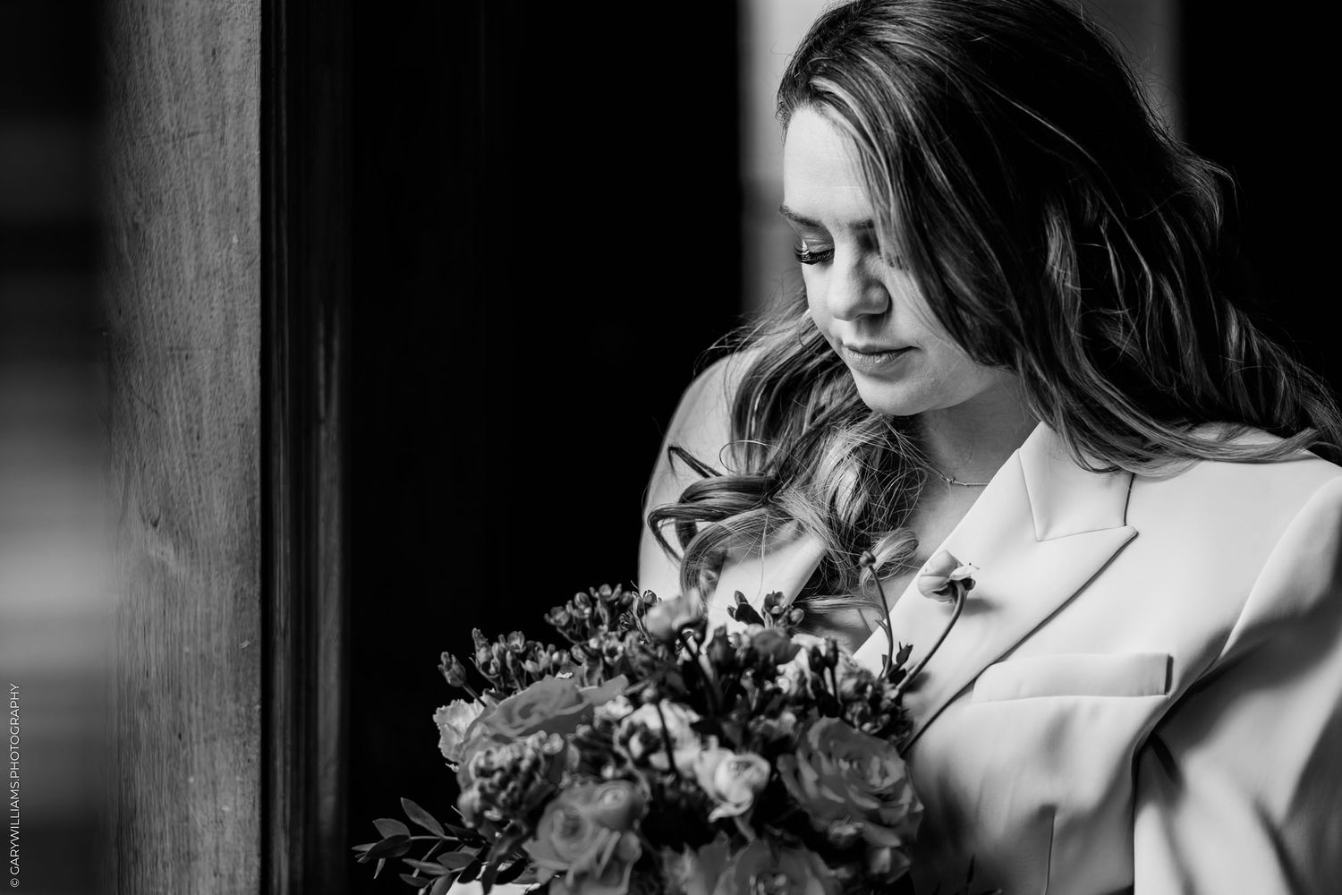 Black and white photo of a bride holding a bouquet of flowers, looking down thoughtfully near a wooden pillar.