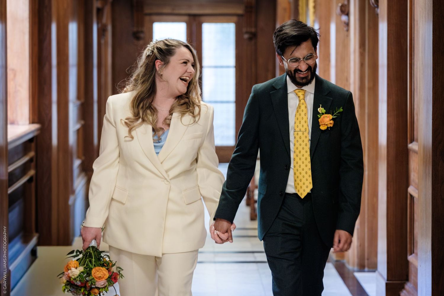 Happy couple in wedding attire walking hand-in-hand, bride in cream suit holding bouquet, groom in dark suit with yellow tie.