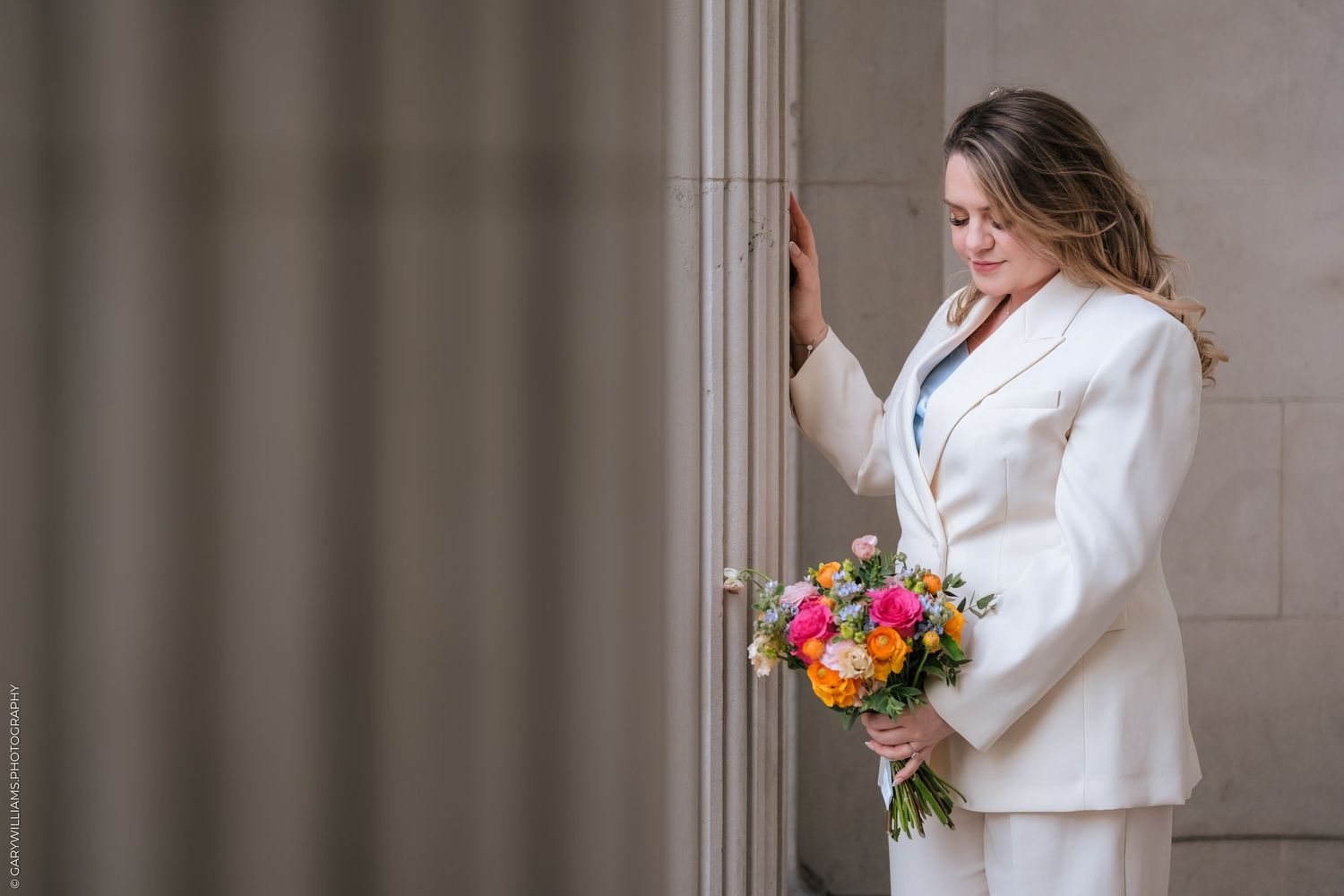 Bride in white suit holding colorful bouquet, leaning against stone column in elegant wedding portrait.