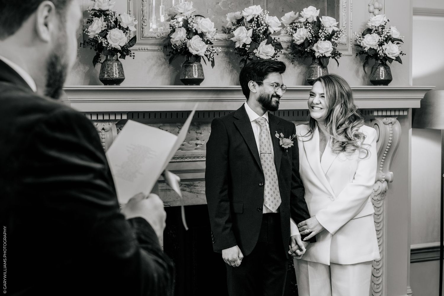 Black and white wedding ceremony photo of couple exchanging vows before officiant, with floral arrangements on mantelpiece.