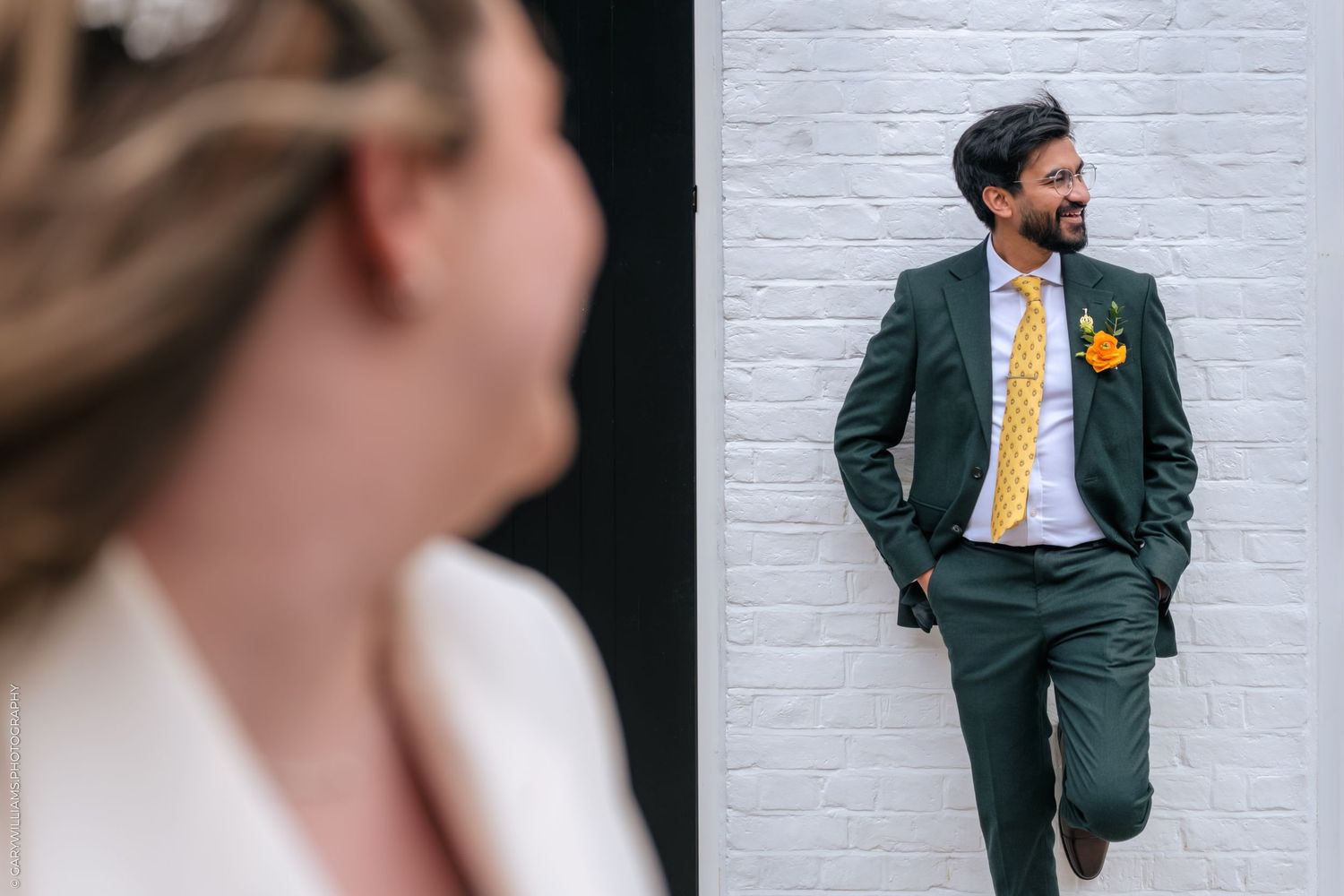 Groom in green suit with yellow tie and floral boutonniere leans against white brick wall, bride in white blurred foreground.