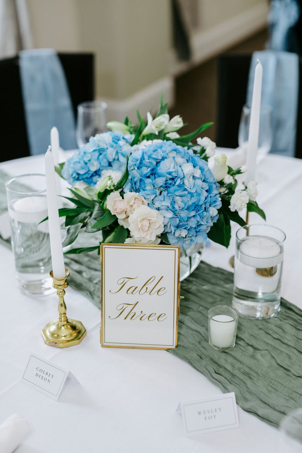 Blue hydrangea floral centerpiece with white candles and gold Table Three sign on white linen at wedding reception.