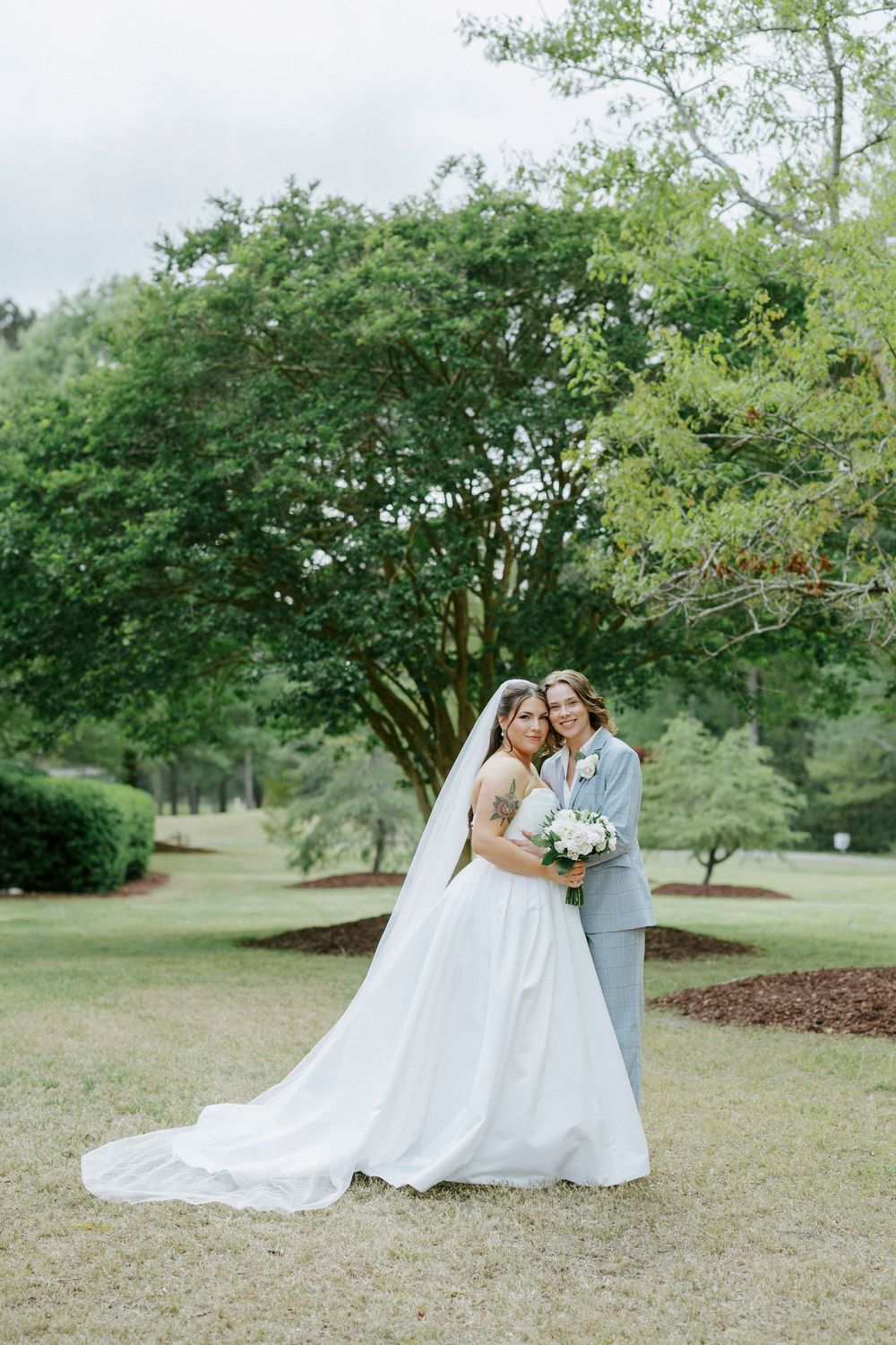 Two brides pose in a lush green park, one in a white gown with veil, the other in a light gray suit.
