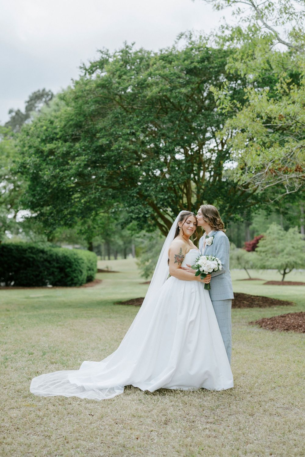 Bride in white gown with veil and groom in light gray suit embrace outdoors in lush green garden setting.