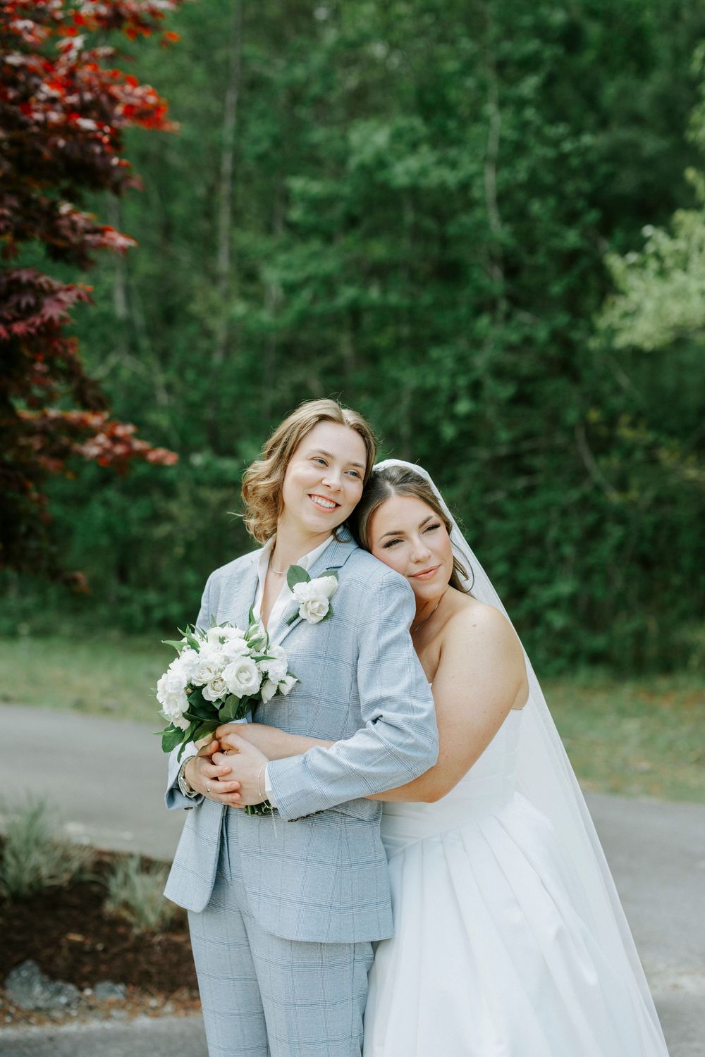 Two brides pose together outdoors, one in white wedding dress with veil, other in light blue suit holding bouquet.