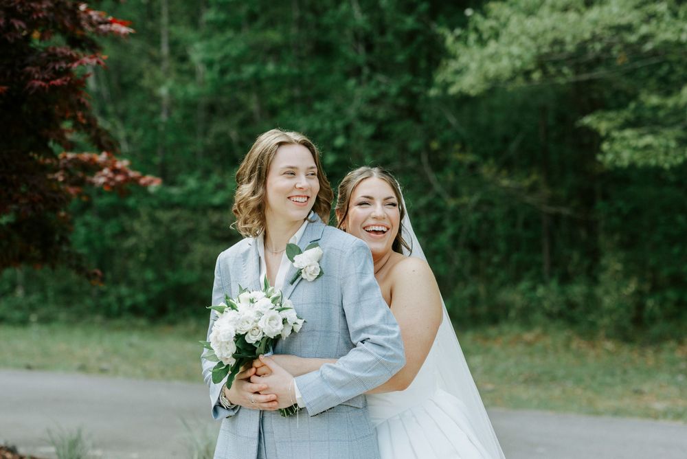 Two brides pose together outdoors, one in a gray suit holding a white bouquet, the other in a white wedding gown, smiling joyfully.