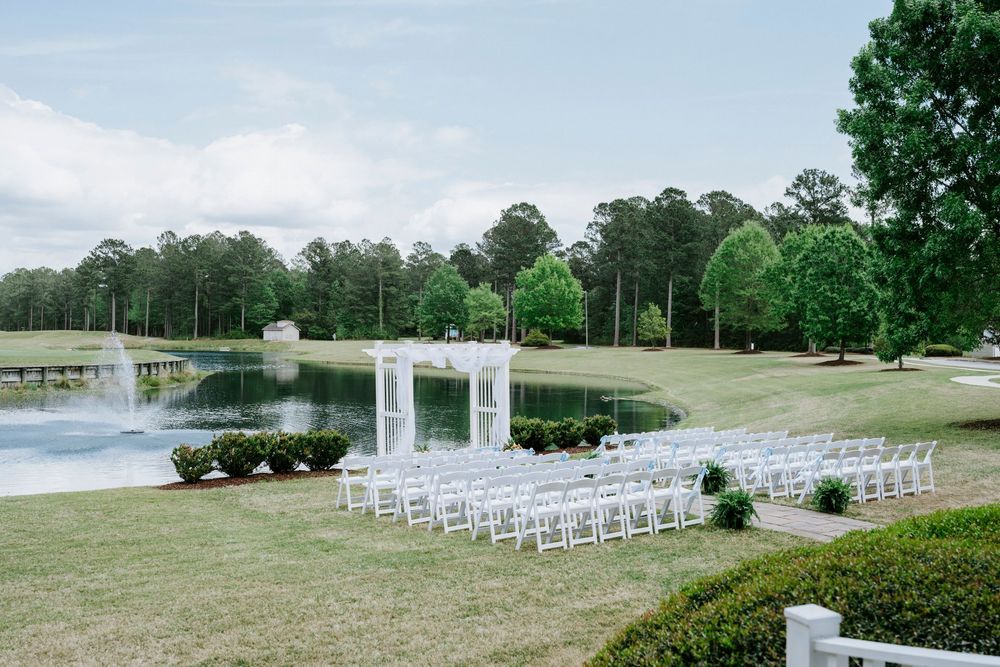 Elegant outdoor wedding ceremony setup with white chairs facing lakeside altar amid lush greenery.