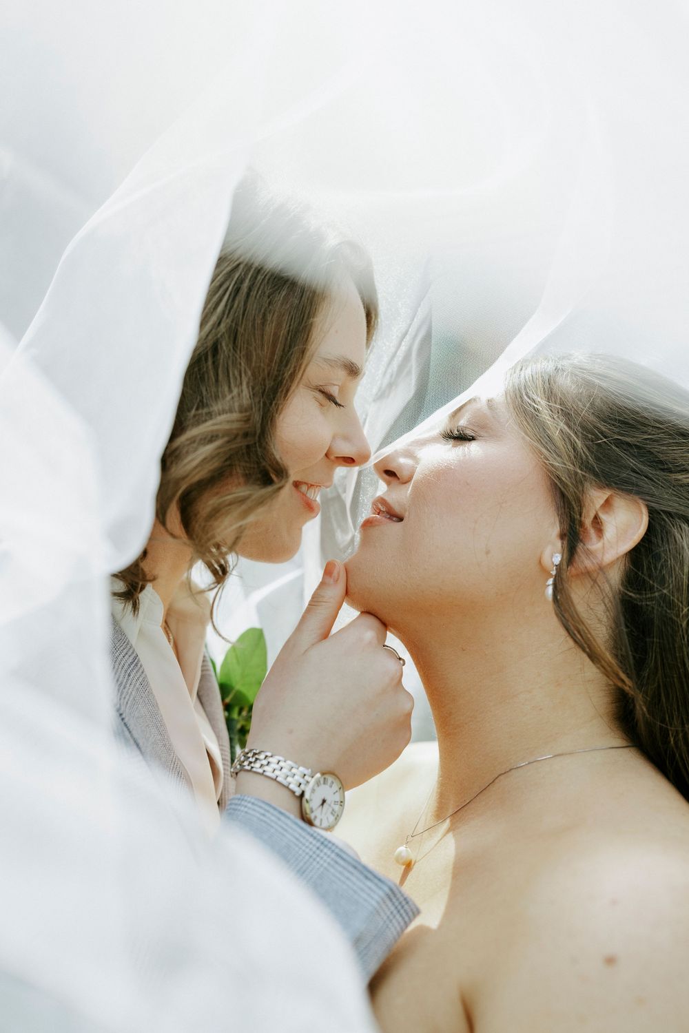 Two brides share a tender moment under a flowing white veil, nearly kissing in a romantic wedding photo.