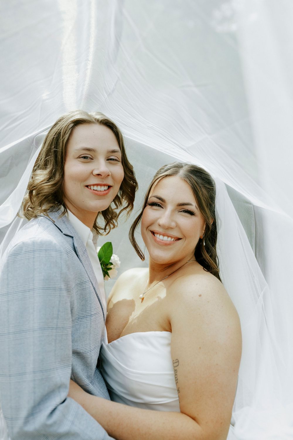 Two brides smile under a flowing white veil, one in a gray suit with boutonniere, one in a strapless white dress.