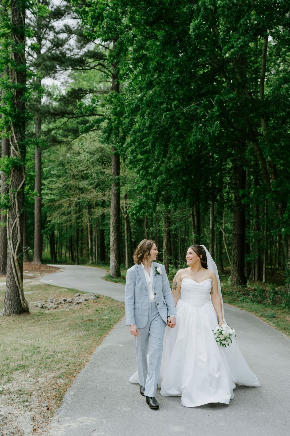 Two brides walk hand-in-hand down a tree-lined path, one in a gray suit, one in a white ball gown with veil.