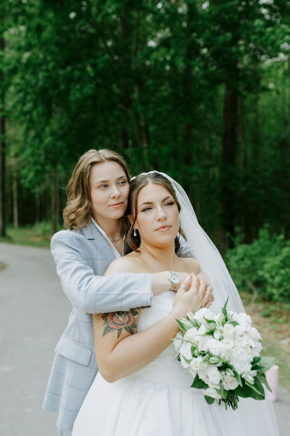 Two brides embrace on tree-lined path, one in white gown holding white bouquet, other in gray suit with tattoo.