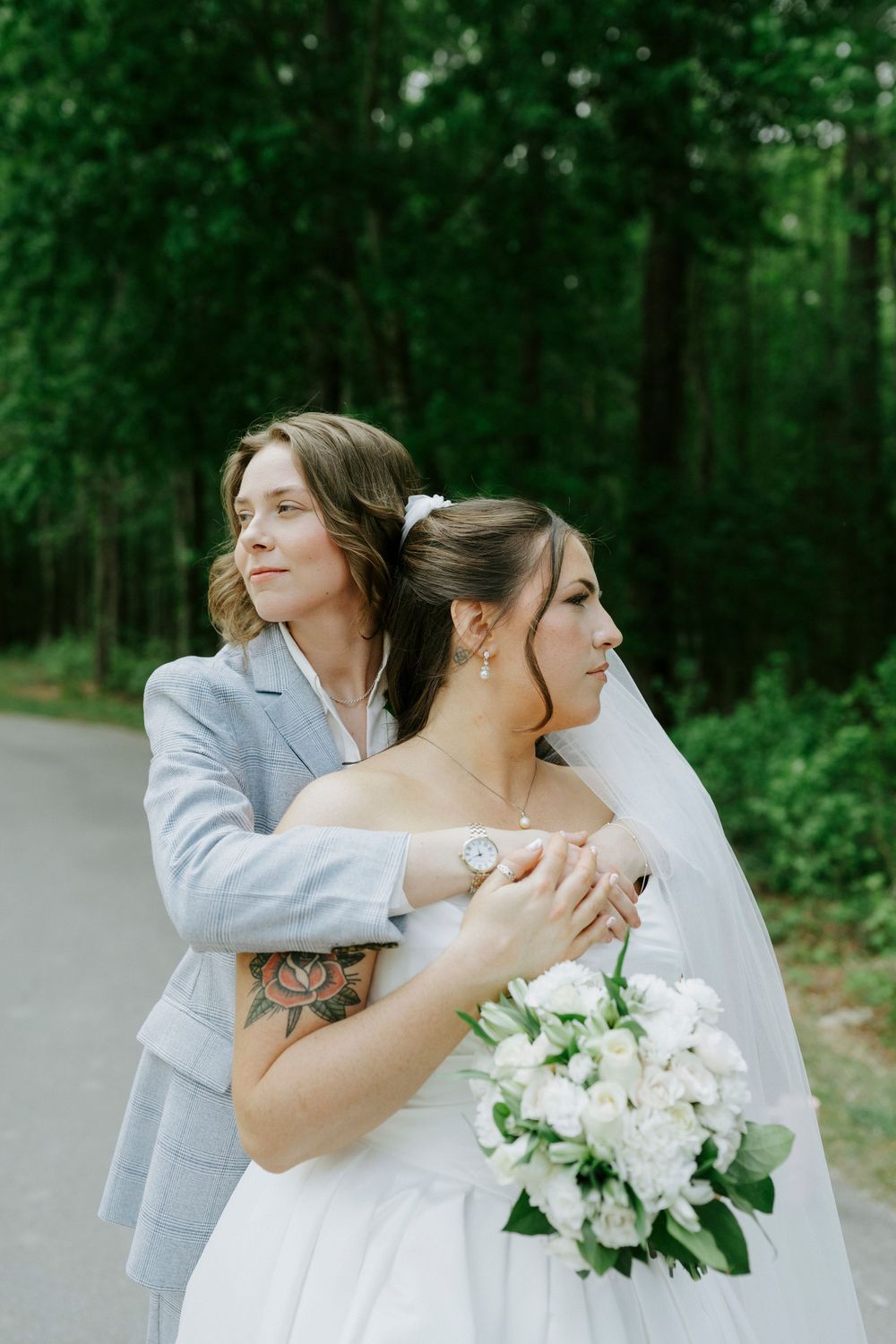A bride in a gray suit embraces her partner in a white strapless gown and veil along a forest path.