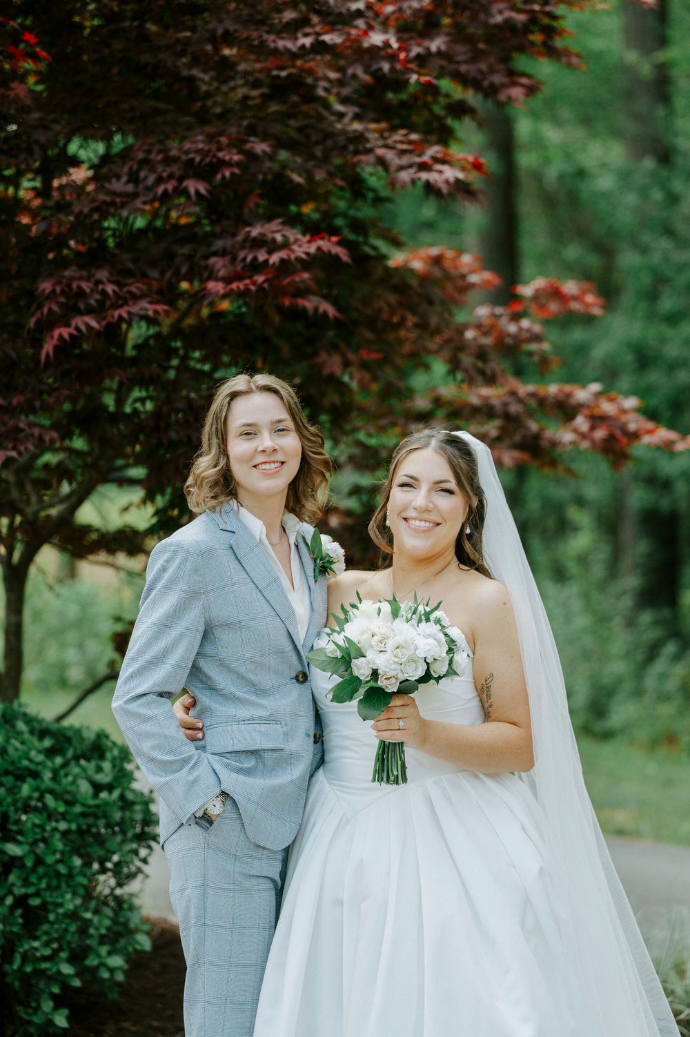 Two brides pose smiling before red maple trees, one in a gray suit, one in a white gown holding white roses.