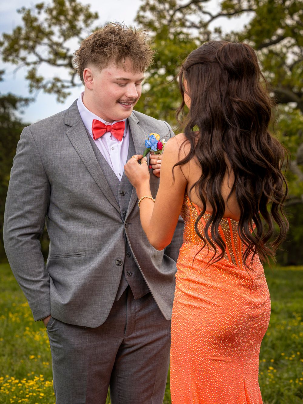 A young couple in prom attire, she pins a boutonniere on his gray suit as he smiles in a green field.