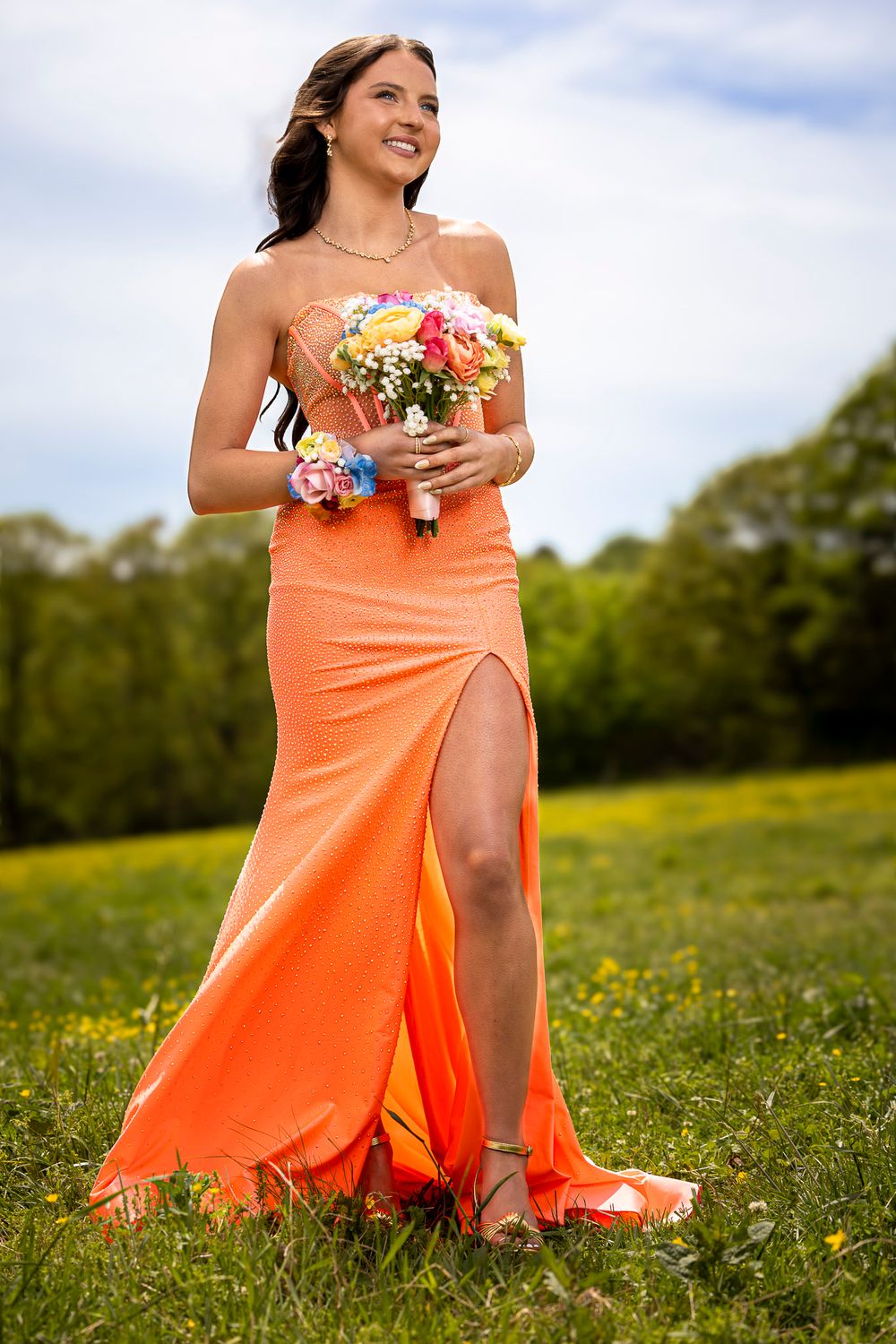 Smiling woman in orange satin gown holding colorful bouquet standing in green meadow.