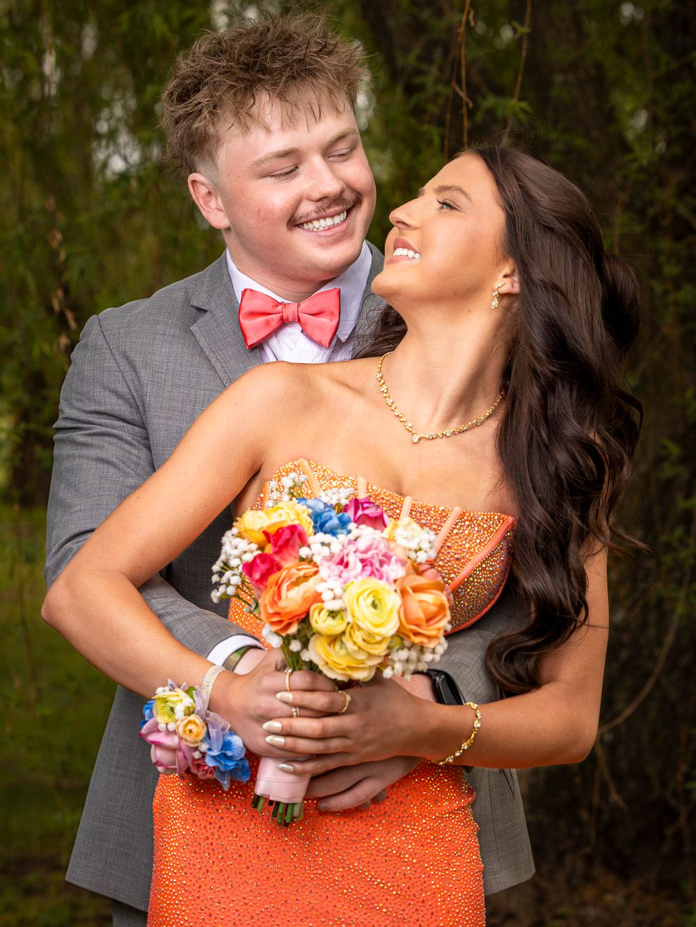 A smiling prom couple embraces outdoors, she holds a colorful bouquet in an orange dress, he wears a gray suit.