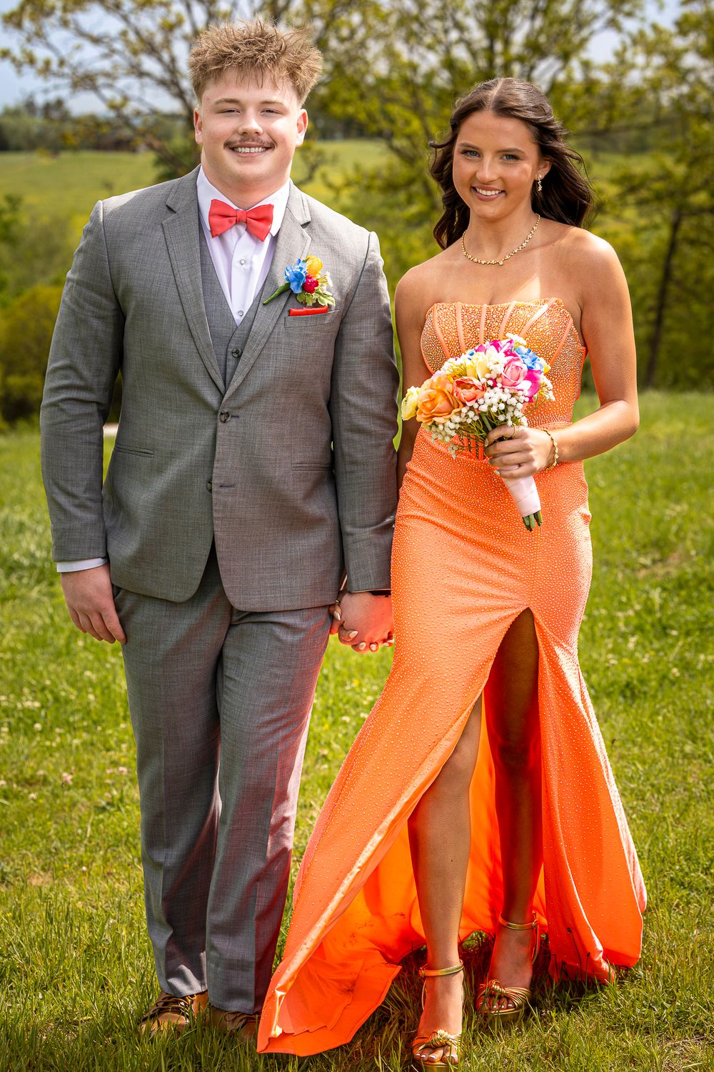 A smiling couple in prom attire, man in gray suit with bow tie, woman in orange gown holding colorful bouquet outdoors.