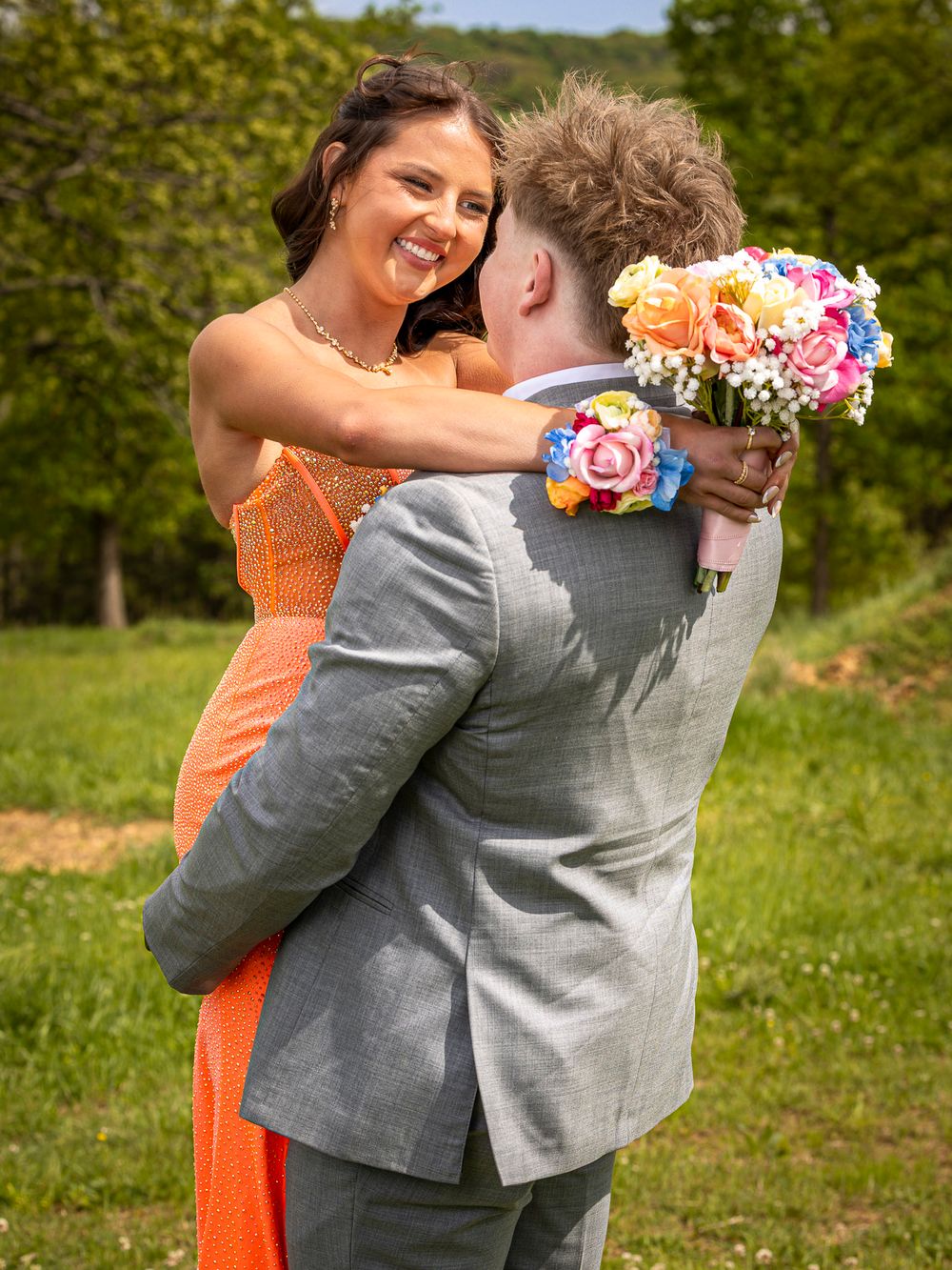 A young woman in an orange prom dress smiles while hugging her date in a gray suit outdoors.